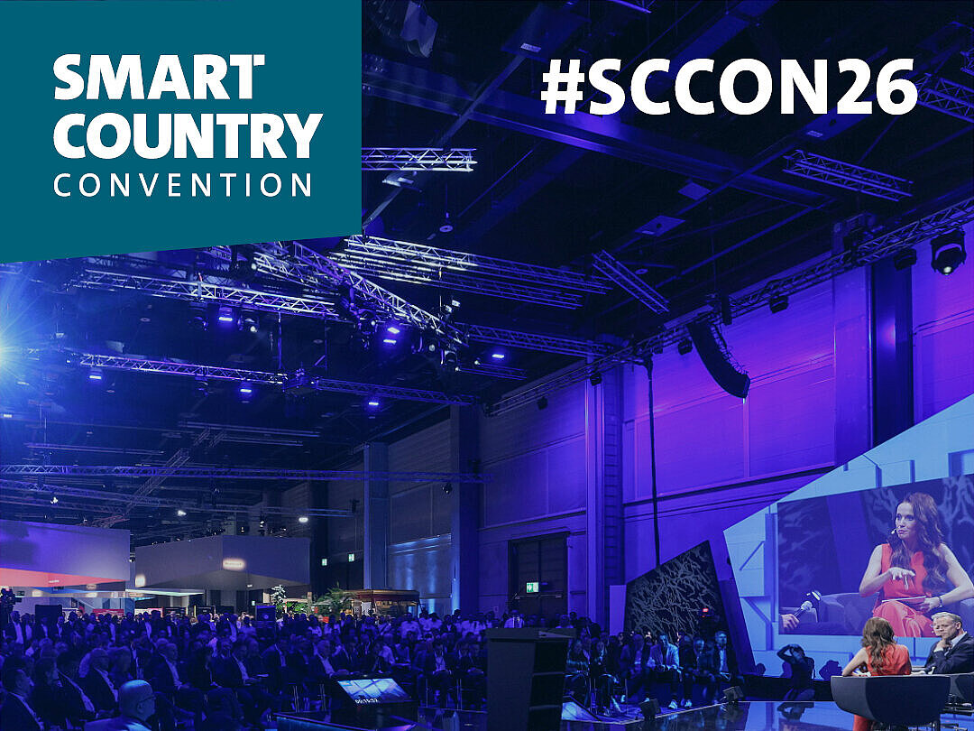 Foto eines Kongresssaales mimt Bühne und Bestuhlung, in lila-blau ausgeleuchtet. Text auf dem Bild in weiß: Smart Country Convention. Save the Date 13.-15. Oktober 2026, Messe Berlin. #SCCON26